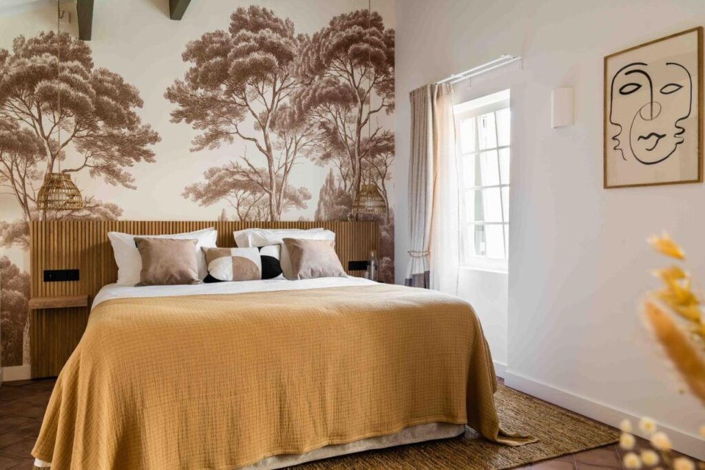 Unique Boutique Hotels in Menorca 30 Cozy bedroom with a large bed, mustard-yellow bedspread, neutral pillows, tree-patterned accent wall, wall art, and wicker lamps—like the inviting rooms found in boutique hotels in Menorca—sunlight streaming through light curtains.