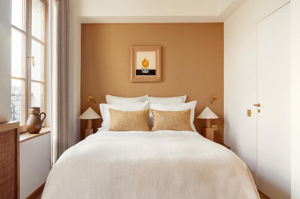 13 Unique Hotels in Paris for a Stylish Stay 26 A cozy bedroom in one of the unique hotels in Paris, featuring a double bed with white linens and beige accent pillows, two bedside tables with lamps, a window with curtains, and framed artwork above the bed on a warm beige accent wall.
