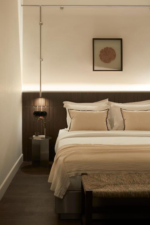 A modern, minimal bedroom inspired by boutique hotels in Athens, with a neatly made bed in beige and white bedding, a dark wooden headboard, a bedside table with a lamp, a woven bench at the foot, and framed artwork on the wall.