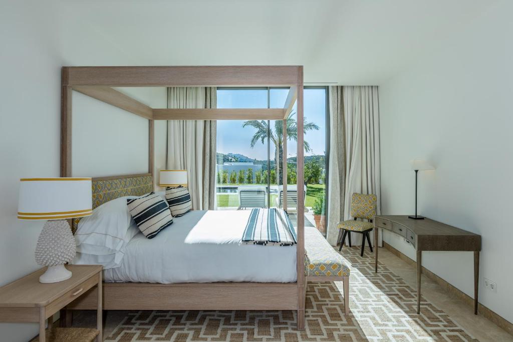 TOP 10 Luxury Hotels in Andalucía 8 Bright, modern bedroom with a wooden canopy bed, striped pillows, and a patterned rug. Reminiscent of luxury hotels in Andalucía, the space features a large garden-view window and minimalist decor with a desk, chair, nightstand, and lamp.