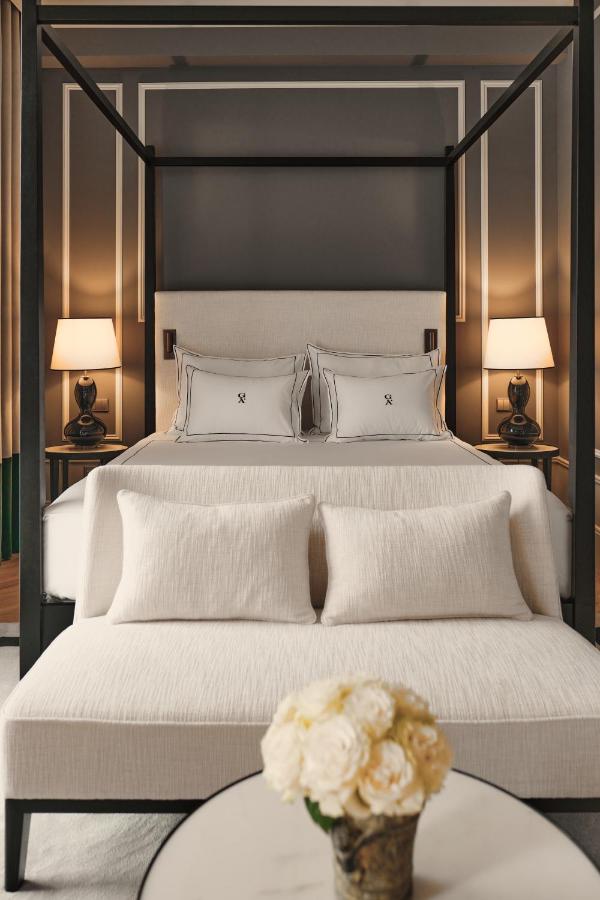 Top 12 Boutique Hotels in Porto, Portugal 4 A modern bedroom reminiscent of boutique hotels in Porto, featuring a four-poster bed with cream bedding, a matching bench, bedside tables with lamps, and a round table adorned with a vase of white roses.