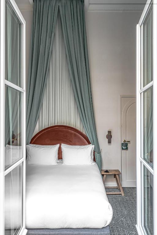 13 Unique Hotels in Paris for a Stylish Stay 53 A cozy bedroom in one of the unique hotels in Paris, featuring a double bed with white bedding, a curved brown headboard, tall green curtains, a wooden stool, and a small door with a Do Not Disturb sign—all viewed through glass doors.