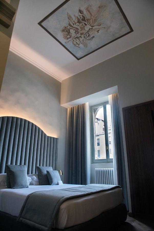 13 UNIQUE Boutique Hotels in Rome 12 A stylish bedroom inspired by boutique hotels in Rome, Italy, features a tall, curved upholstered headboard, blue-gray bedding and pillows, a large window with sheer curtains, and decorative floral artwork on the ceiling.