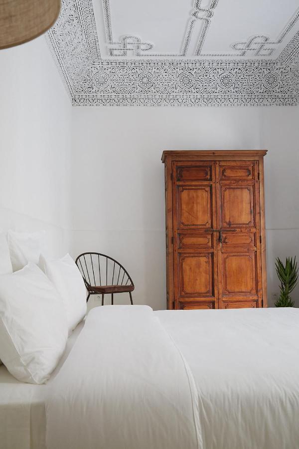 10 Unique Riads in Marrakech with Pool 14 A minimalist bedroom with white walls and bedding, an ornate white ceiling, a wooden wardrobe, a black chair, and a green plant in the corner—reminiscent of the serene elegance found in riads in Marrakech with pool.