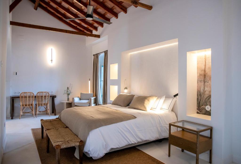 TOP 10 Luxury Hotels in Andalucía 35 Bright, modern bedroom with wooden beams, a double bed with neutral bedding, a woven bench, rattan side table, chair, and desk. Warm lighting and minimal decor evoke the cozy elegance found in luxury hotels in Andalucía.