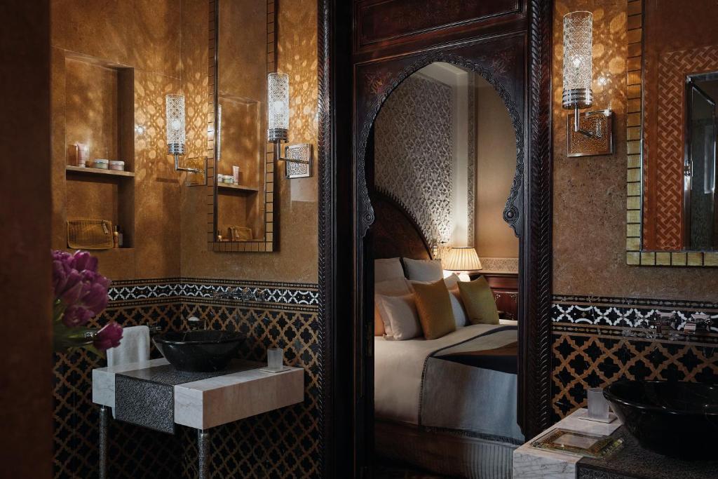 10 Unique Riads in Marrakech with Pool 38 Elegant bathroom with ornate Moroccan-style decor, patterned tiles, marble sinks, and warm lighting. An arched doorway reveals a cozy bedroom—echoing the luxurious charm found in riads in Marrakech with pool.