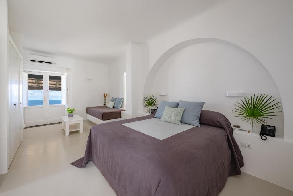 7 Best Hotels in the Aeolian Islands 5 A bright, minimalist bedroom inspired by the best hotels in the Aeolian Islands, with a large bed draped in gray and blue linens, a small sofa, white walls, a table with green plants, and glass doors flooding the space with natural light.