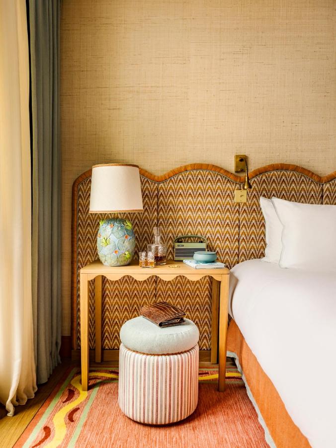 13 Unique Hotels in Paris for a Stylish Stay 5 A cozy hotel room corner, typical of unique hotels in Paris, features a patterned headboard, wooden nightstand with a lamp, books, water glasses on a tray, a round ottoman, and part of a neatly made bed with white linens.
