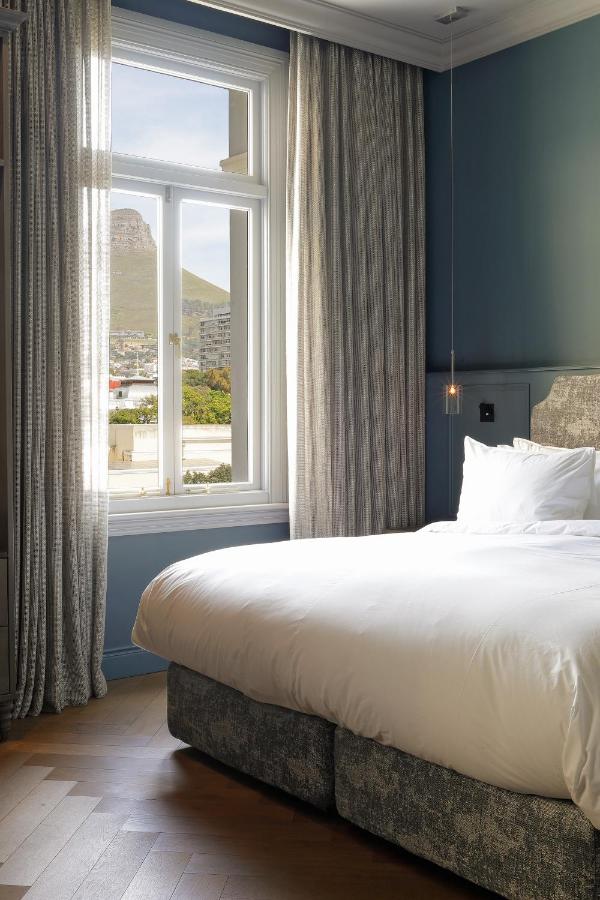Unique Places to Stay in Cape Town 17 A neatly made bed with white linens sits by a large window framed with gray curtains. Blue walls and wooden floors complete this serene space—one of the unique places to stay in Cape Town, offering mountain and city views under a clear sky.