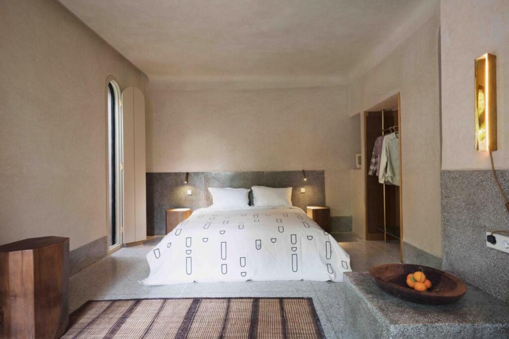 10 Unique Riads in Marrakech with Pool 10 Minimalist bedroom inspired by riads in Marrakech with pool, featuring a stone platform bed, white geometric bedding, built-in nightstands, soft beige walls, an open closet with hanging clothes, a bowl of fruit, and arched shuttered windows.