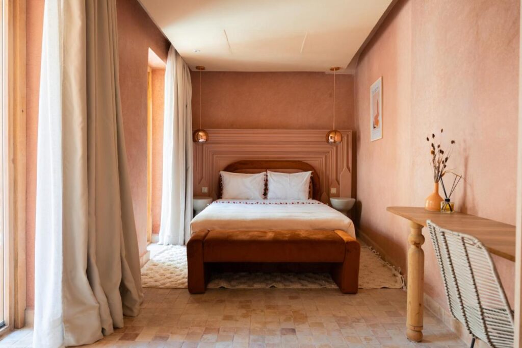 10 Unique Riads in Marrakech with Pool 26 A cozy bedroom inspired by riads in Marrakech with pool features a double bed, white bedding, tan upholstered headboard and bench, two hanging lamps, light pink walls, a wooden desk with chair, and light curtains by the window.