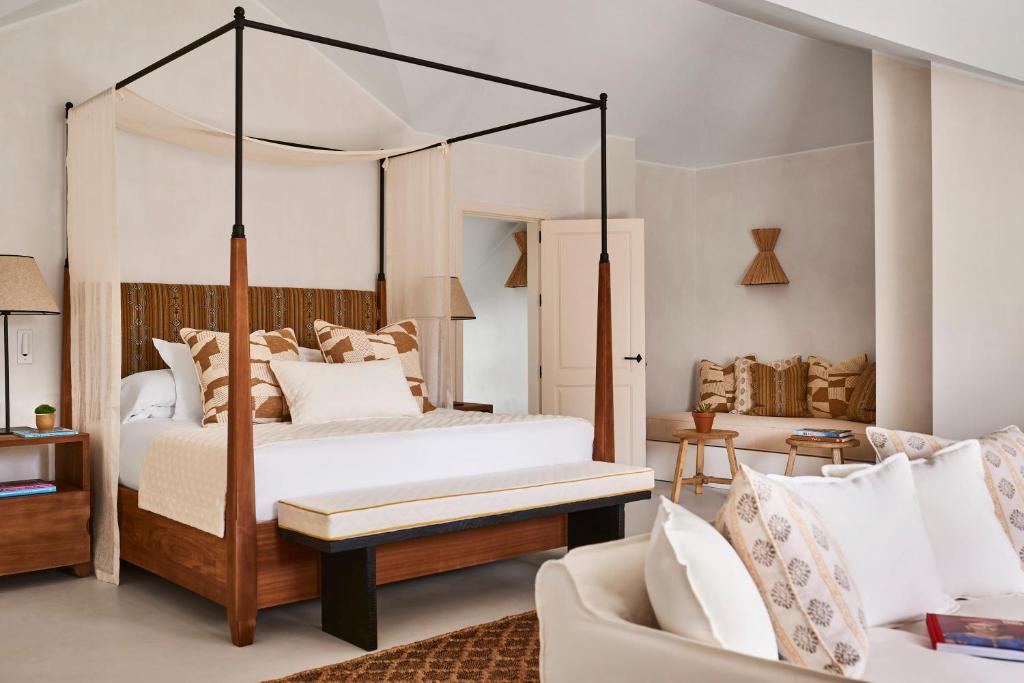 TOP 10 Luxury Hotels in Andalucía 18 A stylish bedroom inspired by luxury hotels in Andalucía, featuring a four-poster bed, neutral-toned bedding, patterned pillows, a wooden bench, cozy seating with cushions, and warm natural lighting. The decor blends modern style with earthy, rustic elements.