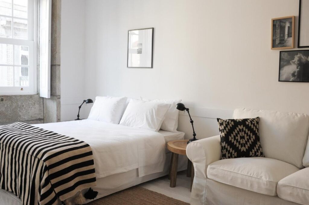 Top 12 Boutique Hotels in Porto, Portugal 44 A bright, minimal bedroom inspired by boutique hotels in Porto, featuring a double bed with white bedding, a striped blanket, a white armchair with a patterned cushion, wall art, a small wooden side table, and large windows flooding the space with natural light.