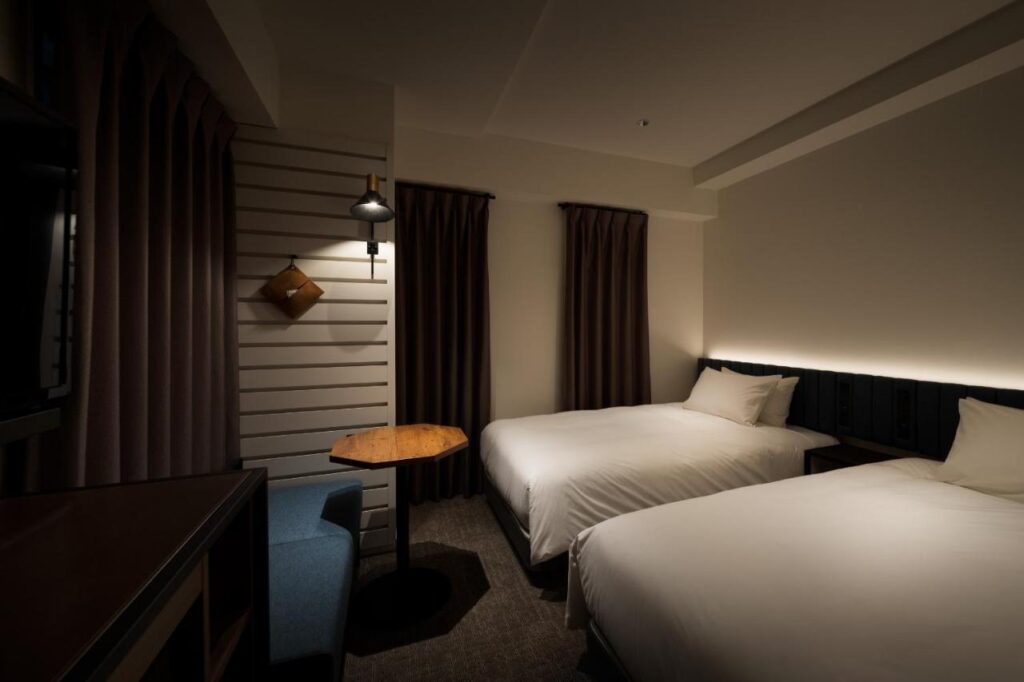 7 Best Hotels in Hiroshima, Japan 17 A modern hotel room with two neatly made single beds, a blue chair by a wooden table, and brown curtains. Soft lighting creates a cozy ambiance, making it a perfect choice among the best hotels in Hiroshima Japan.