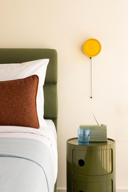 A modern bedroom reminiscent of boutique hotels in Athens features a green upholstered headboard, a white pillow with a brown cushion, a round green bedside table with a retro radio, a glass of water, and a yellow wall light with pull chain.