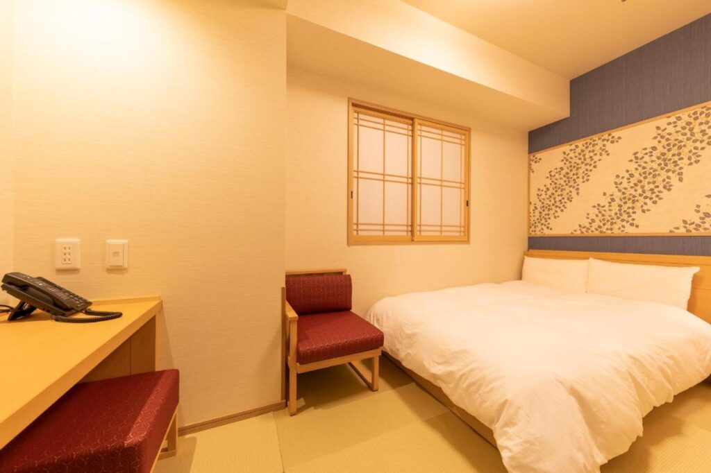 15 Unique Hotels in Tokyo: Coolest hotels 17 A cozy hotel room in one of the unique hotels in Tokyo, featuring a neatly made double bed, a wooden desk with a telephone, a cushioned chair, and a wood-framed window with rice paper. Soft lighting and floral wall art add to the inviting decor.