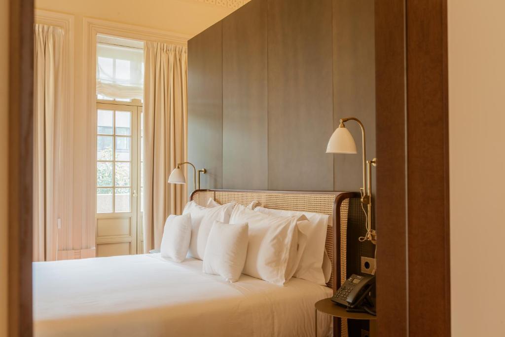 Top 12 Boutique Hotels in Porto, Portugal 48 A neatly made bed with white linens and multiple pillows sits against a tall wooden headboard, reflecting the elegance of boutique hotels in Porto. Light streams in through large windows with cream-colored curtains above bedside tables.