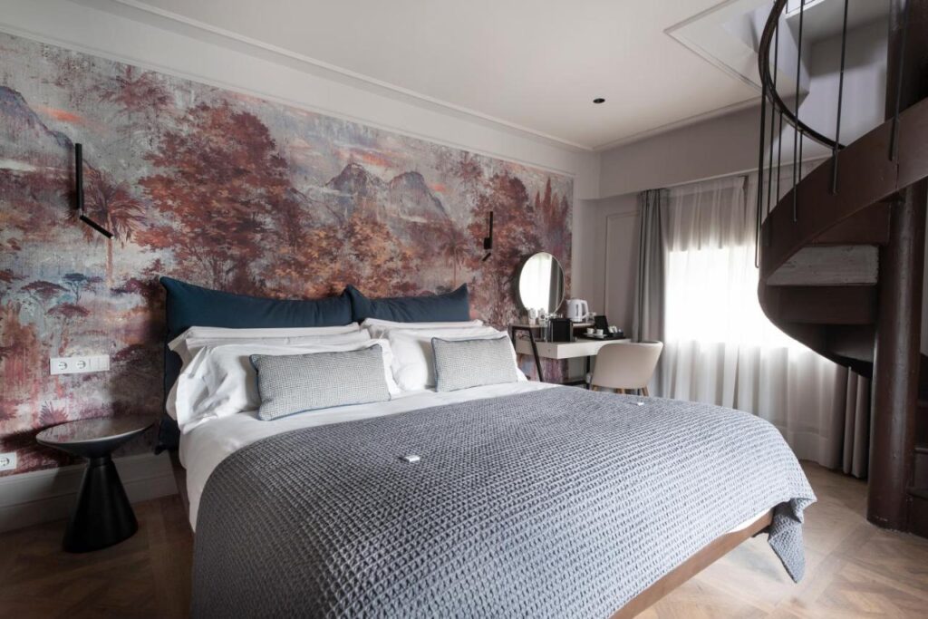A modern bedroom inspired by hotels in Cantabria, featuring a double bed with blue-gray bedding, a scenic mural, a desk with mirror, kettle, white chair, and a spiral staircase near a window dressed in sheer curtains.