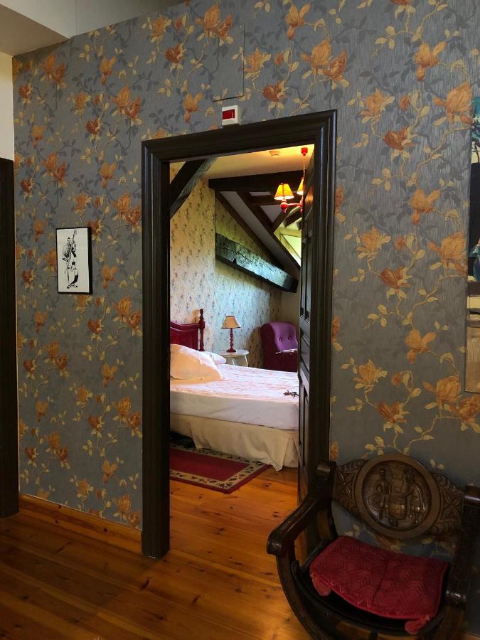 A doorway opens from a hallway with floral wallpaper into a cozy bedroom reminiscent of charming hotels in Cantabria, featuring a bed, vintage lamp, armchair, and wooden floors. A carved wooden chair sits near the doorway, adding to the warm, classic decor.