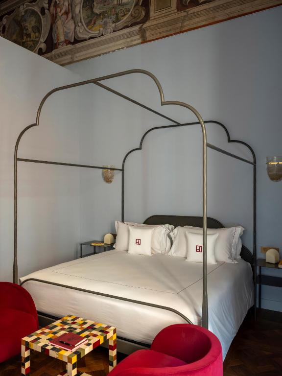 13 UNIQUE Boutique Hotels in Rome 50 A stylish bedroom reminiscent of boutique hotels in Rome, featuring a metal four-poster bed, white bedding, two red armchairs, a colorful checkered table, and ornate ceiling frescoes above pale blue walls.