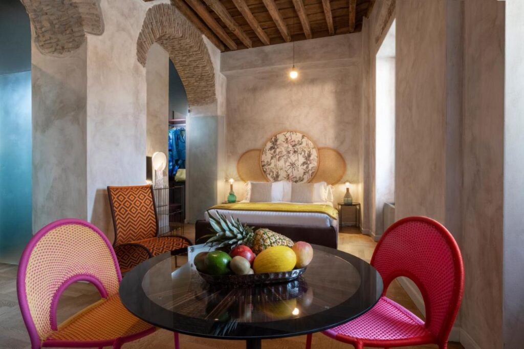 13 UNIQUE Boutique Hotels in Rome 39 A stylish bedroom inspired by boutique hotels in Rome, with rustic stone walls and ceiling, a bed with a floral headboard, colorful chairs around a glass table, and a fruit bowl centerpiece. Warm lighting and eclectic decor create a cozy atmosphere.