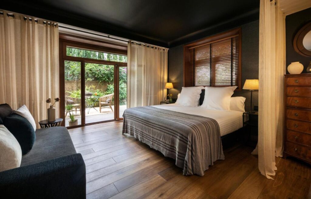 13 Unique Hotels in Barcelona, Spain 11 A cozy bedroom found in unique hotels in Barcelona, featuring a large bed, neutral bedding, bedside lamps, sheer curtains, wood floors, a dark sofa, and glass doors opening to a patio with outdoor furniture and lush greenery.