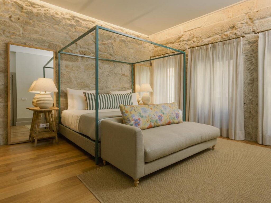 Top 12 Boutique Hotels in Porto, Portugal 24 A cozy bedroom reminiscent of boutique hotels in Porto, with a four-poster bed, striped pillows, a floral cushion on a beige bench, stone walls, wood floor, sheer white curtains, and two lamps on wooden side tables.