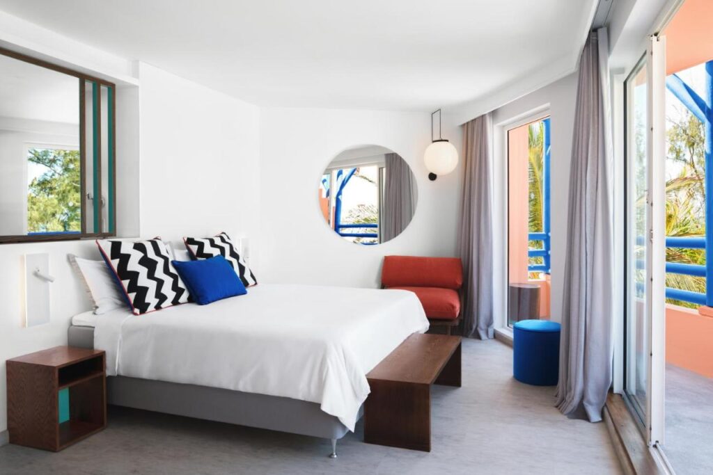 Most Unique Adults Only Hotels in Mauritius, Indian Ocean 5 A modern adults only hotel room in Mauritius with a large bed, geometric-patterned pillows, a round mirror, an orange armchair, and a balcony with blue railings and sheer curtains letting in natural light.