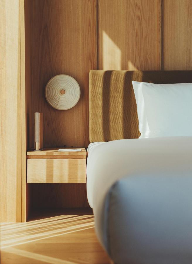 15 Unique Hotels in Tokyo: Coolest hotels 3 A close-up of a modern, minimalist bedroom at one of the unique hotels in Tokyo, featuring a bed with white linens, a wooden headboard, a small nightstand, and a round wall-mounted light fixture against light wood-paneled walls.
