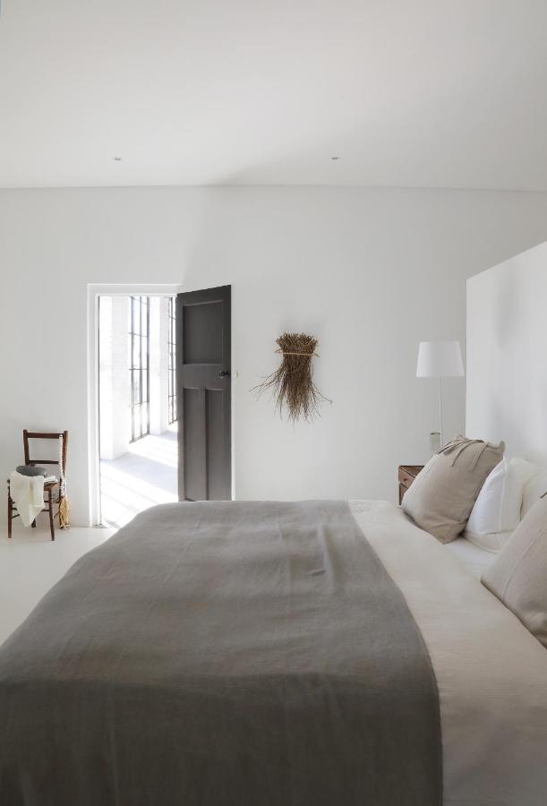 Unique Places to Stay in Cape Town 25 Minimalist bedroom in neutral tones, featuring a wooden chair with a draped throw and dried plants on the wall—one of the unique places to stay in Cape Town, where natural light streams through an open door.