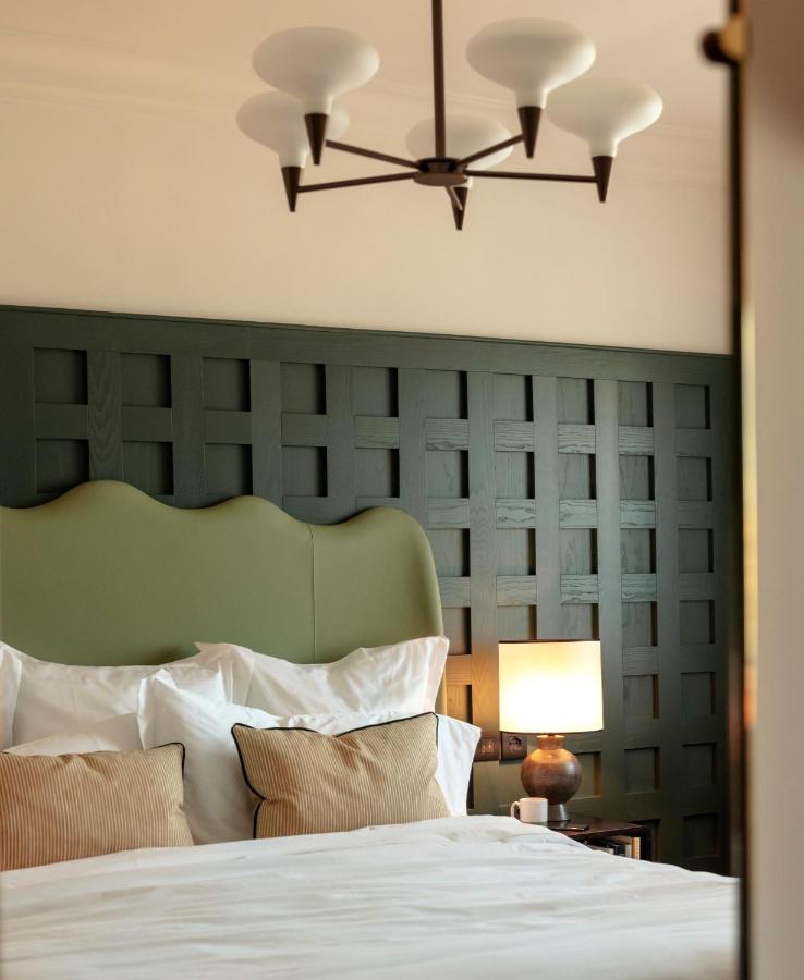 13 UNIQUE Boutique Hotels in Rome 24 A neatly made bed with white linens and beige pillows sits in front of a dark green, lattice-patterned headboard—evoking the refined charm found in boutique hotels in Rome, Italy. A bedside table holds a lit lamp and cup, with a modern chandelier above.