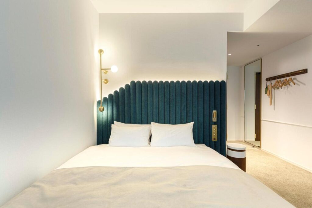 7 Best Hotels in Hiroshima, Japan 10 A modern hotel room at one of the best hotels in Hiroshima, Japan, featuring a double bed with a teal upholstered headboard, crisp white bedding, gold reading lights, a small nightstand, and a handy coat rack near the doorway.