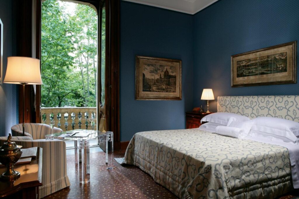 13 UNIQUE Boutique Hotels in Rome 23 Elegant bedroom with blue walls, a patterned bedspread, classic furniture, and framed artwork—reminiscent of the sophisticated style found in boutique hotels in Rome, Italy—plus a balcony offering views of lush green trees.