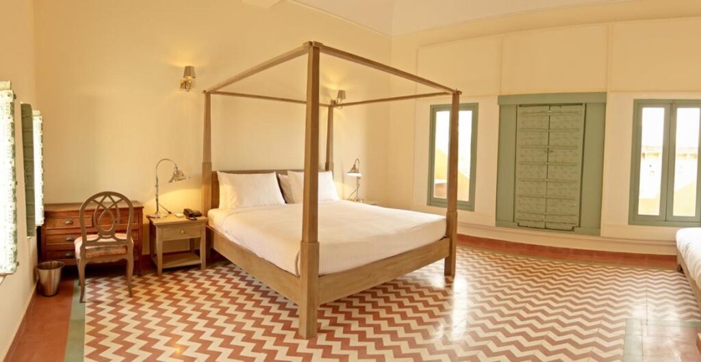 Authentic Heritage Hotels in Mandawa 3 A spacious, well-lit bedroom in the style of hotels in Mandawa, featuring a wooden four-poster bed, bedside table with a lamp, wooden desk and chair, large windows, and a red and white zigzag patterned floor.