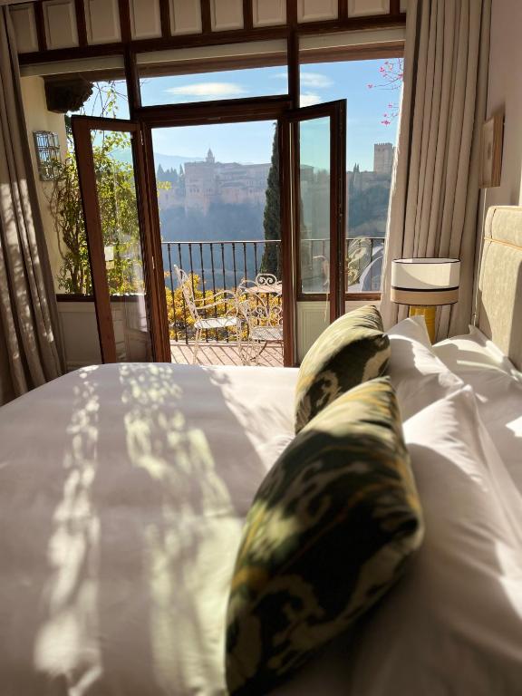 Sunlit bedroom with white bedding and patterned pillows, open glass doors leading to a balcony with a table and chairs, and a scenic view of a castle—perfect for those seeking boutique hotels in Granada.