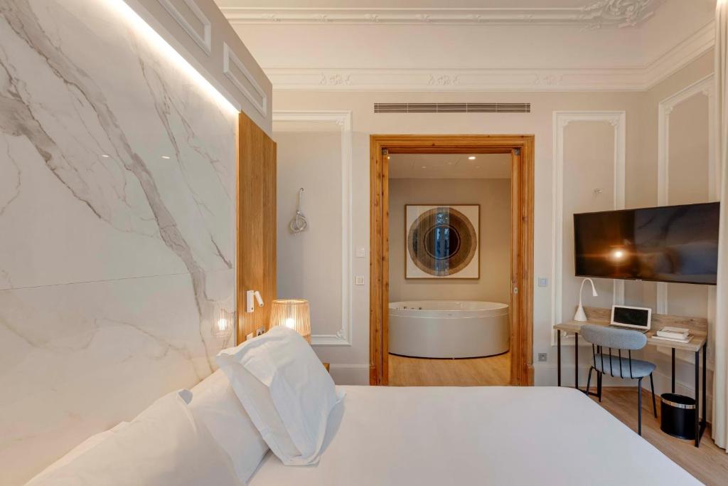 13 Unique Hotels in Barcelona, Spain 35 A modern, elegant hotel room—one of the unique hotels in Barcelona—features a large bed, white linens, a marble accent wall, flat-screen TV, desk with lamp and laptop, and an open doorway leading to a round bathtub in the adjacent room.