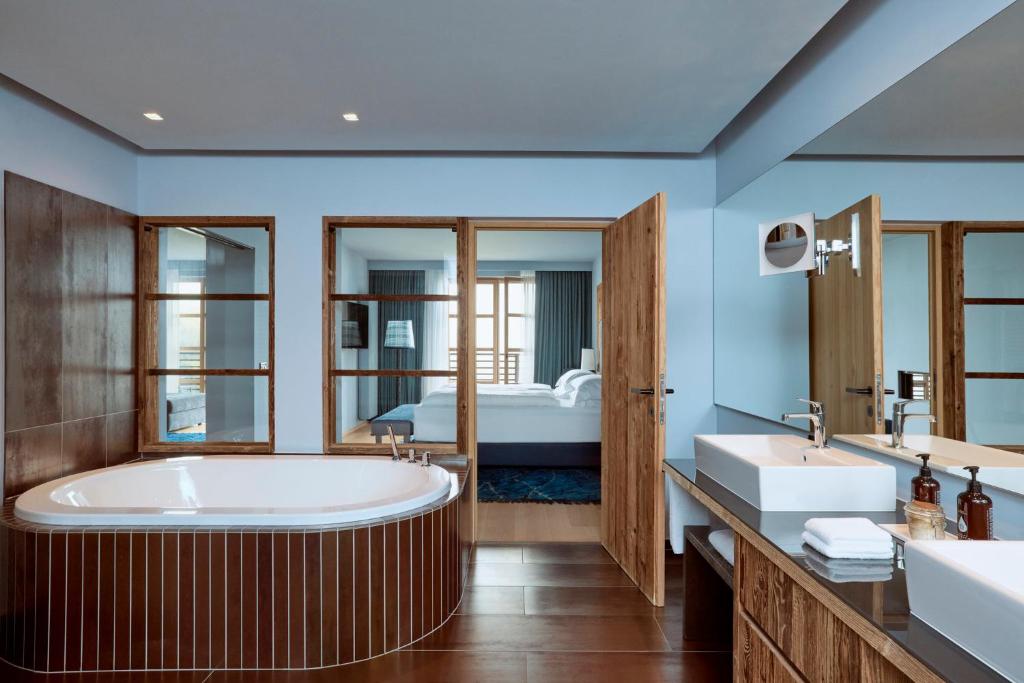 Modern bathroom with a large oval bathtub, double sinks, wooden accents, and glass doors opening to a bedroom with a neatly made bed and blue curtains—typical of the luxury you’ll find at the best hotels in the Dolomites.