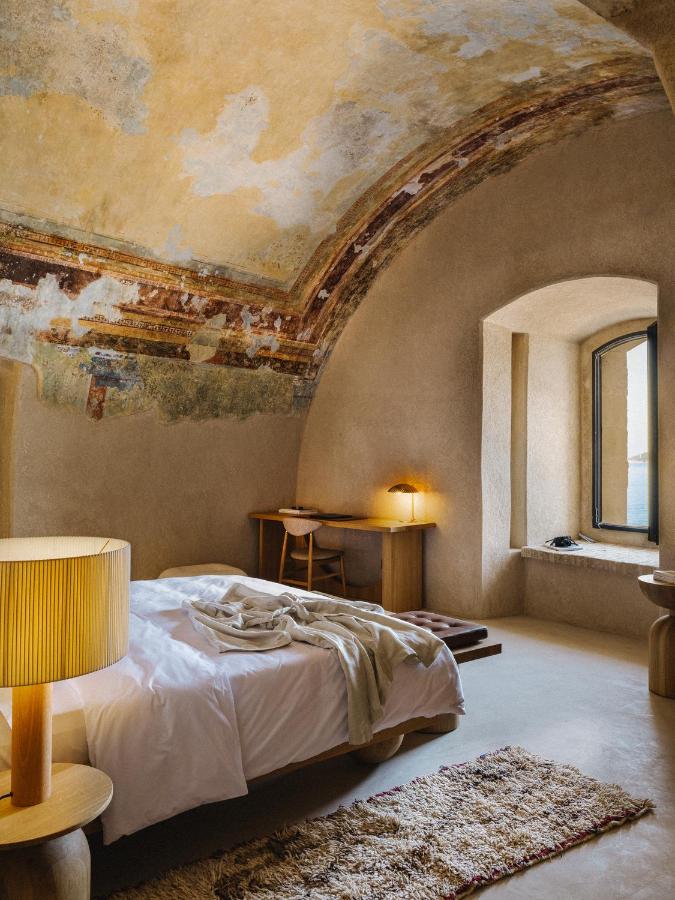 Most Authentic Boutique Hotels in Montenegro, Europe 4 A cozy bedroom inspired by boutique hotels in Montenegro, with rustic, textured walls, a curved ceiling with faded frescoes, a bed with white linens, a wooden desk and chair, a lamp, and an arched window letting in natural light.