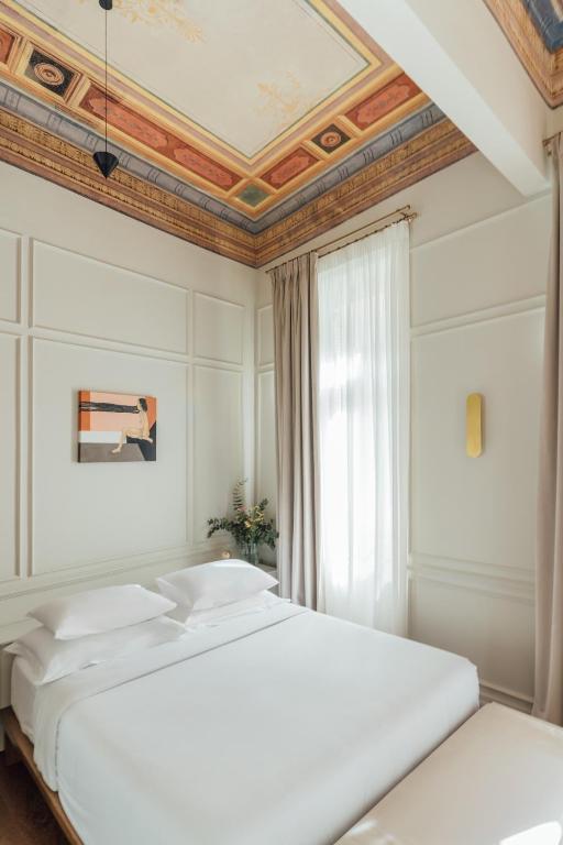 A bright, elegant bedroom reminiscent of boutique hotels in Athens, featuring a white bed, tall window with beige curtains, a modern painting on the wall, ornate ceiling details, and soft natural light filling the room.