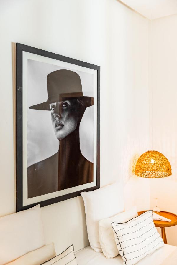 10 Unique Riads in Marrakech with Pool 15 A black-and-white portrait of a person in a wide-brimmed hat hangs above a white couch with cushions; nearby, a wicker lamp glows on a round table, evoking the serene elegance found in riads in Marrakech with pool.