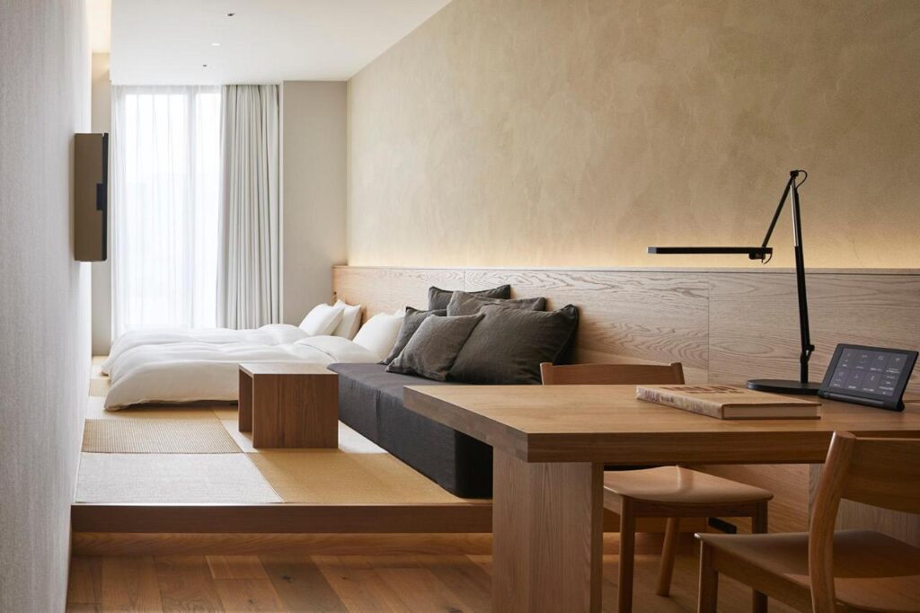 15 Unique Hotels in Tokyo: Coolest hotels 23 A minimalist hotel room—like those found in unique hotels in Tokyo—features low twin beds with white bedding, a black sofa, wooden furniture, a desk with a lamp and tablet, large window with sheer curtains, and soft neutral tones throughout.