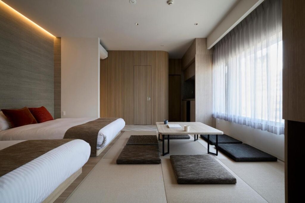 7 Best Hotels in Hiroshima, Japan 8 Minimalist hotel room with two beds, low wooden table, floor cushions, and a large window with sheer curtains—neutral tones and clean lines create a modern, serene atmosphere found in the best hotels in Hiroshima, Japan.