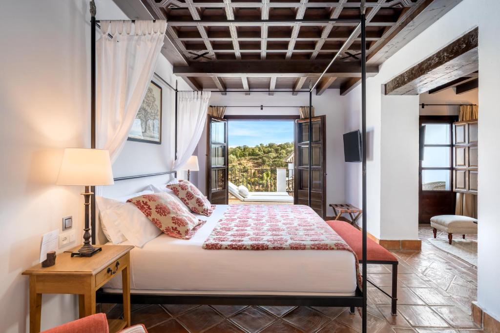 TOP 10 Luxury Hotels in Andalucía 24 A bright bedroom inspired by luxury hotels in Andalucía, featuring a four-poster bed with red and white floral bedding, a wooden ceiling, open French doors to a scenic balcony, and rustic wooden furniture.