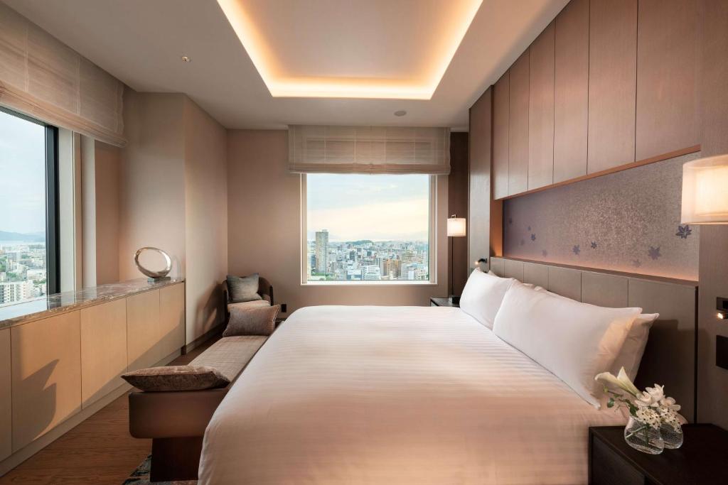 7 Best Hotels in Hiroshima, Japan 18 A modern hotel room at one of the best hotels in Hiroshima features a large bed, neutral decor, wide windows with city views, a built-in bench, and a marble countertop with a round mirror. A vase of white flowers sits on the bedside table.