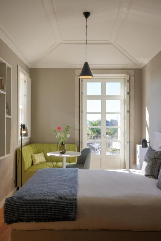 Top 12 Boutique Hotels in Porto, Portugal 31 A bright, modern bedroom inspired by boutique hotels in Porto, featuring a large window, double bed, green corner sofa, small round table with a vase of flowers, and neutral-toned walls with shelves. Sunlight fills the room.