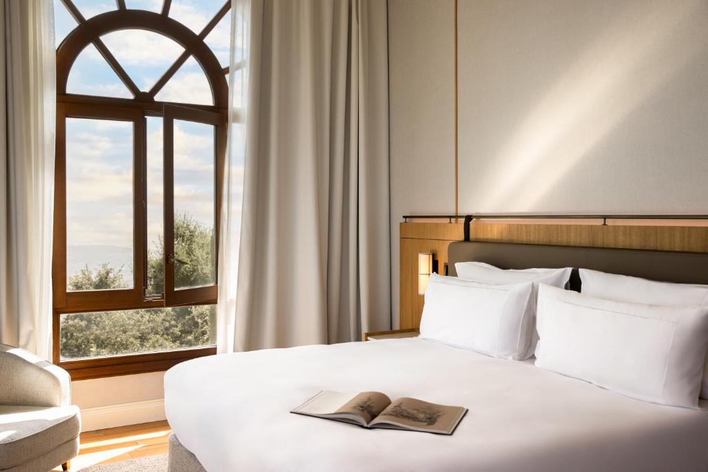 13 Unique Hotels in Barcelona, Spain 49 A neatly made bed with white linens sits beside a large arched window with sheer curtains, letting in natural light—an inviting detail often found in unique hotels in Barcelona. An open book rests on the bed, and a cushioned chair is by the window.