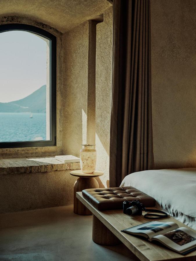 Most Authentic Boutique Hotels in Montenegro, Europe 5 A cozy, sunlit bedroom with beige walls, a bed, and a wooden bench holding a camera and open magazine—evoking the tranquil charm of boutique hotels in Montenegro. A window offers serene lake and mountain views, with a small table and vase nearby.