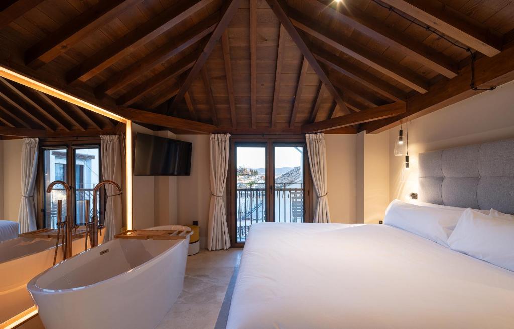 A cozy hotel room with a sloped wooden ceiling, a large bed with white bedding, a freestanding bathtub, and double doors opening to a balcony—offering the charm and comfort found in boutique hotels in Granada.