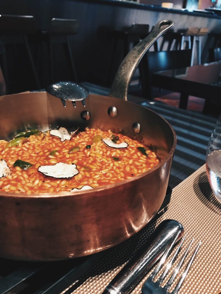 4 Best Hotels in Ribera del Duero, Spain 6 A copper pan filled with creamy risotto topped with truffle slices sits on a table next to a glass of water and silverware, capturing the elegant dining experience often found in top hotels in Ribera del Duero.
