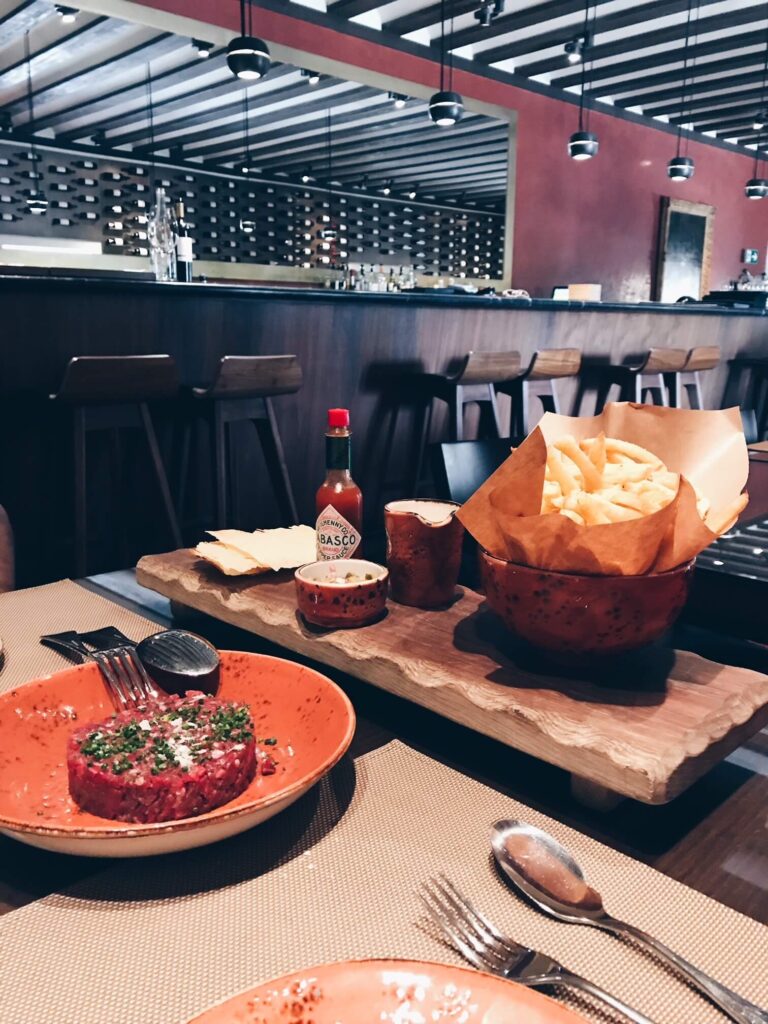 4 Best Hotels in Ribera del Duero, Spain 7 A restaurant table set with steak tartare, fries, Tabasco sauce, bread slices, and cutlery. The modern bar in the background echoes the elegant style often found in top hotels in Ribera del Duero, with high chairs and a wine rack on a red wall.